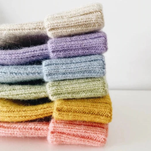 This picture shows a stack of colourful knitted kids winter beanies. In the colours beige, lilac, light blue, lime green, mustard yellow, sorbet pink