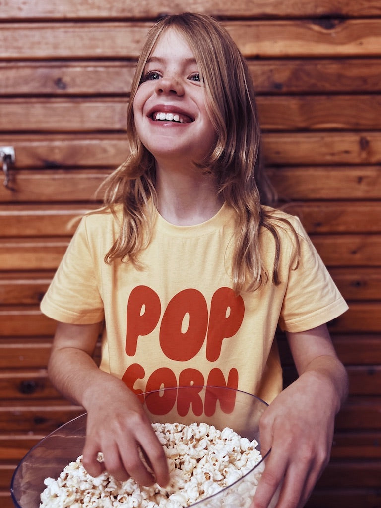This picture shows a girl wearing a yellow cotton t-shirts for kids. With red letters stating popcorn.